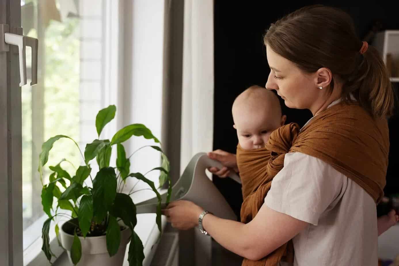madre con bebé en portabebés regando una planta representando la carga mental de la maternidad