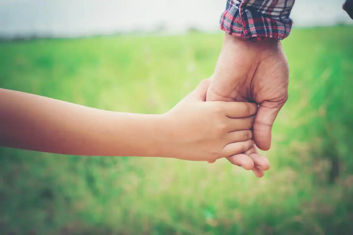Manos de padre e hija entrelazadas en un campo verde, símbolo del vínculo seguro y la autonomía infantil