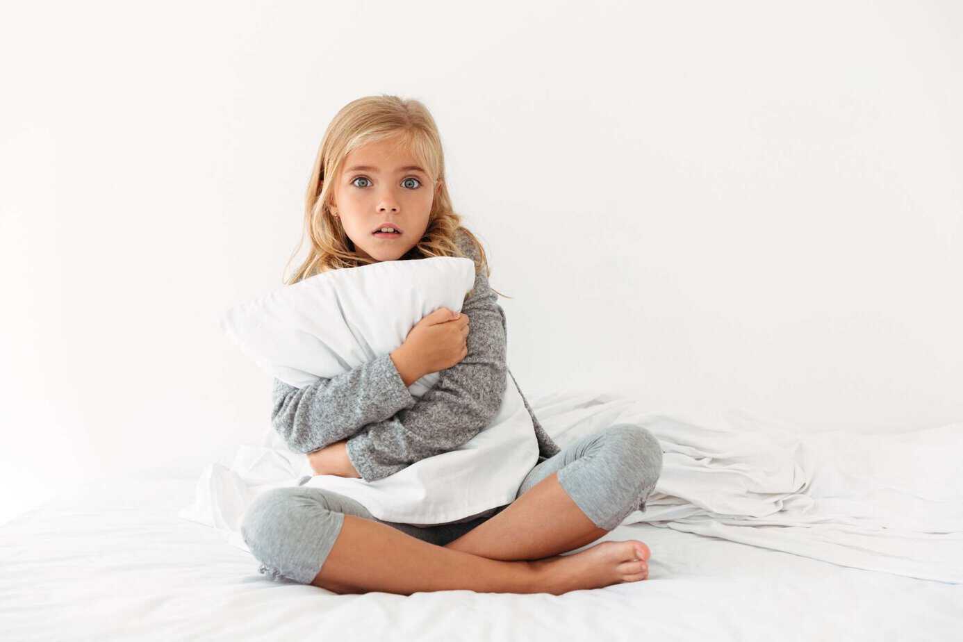 Niña sentada en la cama abrazando una almohada con expresión de miedo, miedos infantiles nocturnos normales