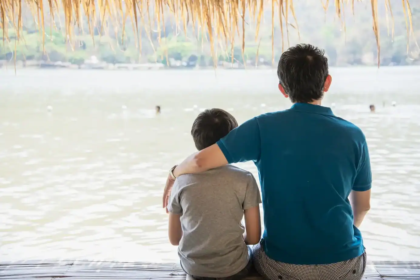 Parentificación emocional. Padre e hijo sentados juntos mirando al agua, con el brazo del padre sobre el hombro del niño, representando al adulto que sigue ocupando su lugar.