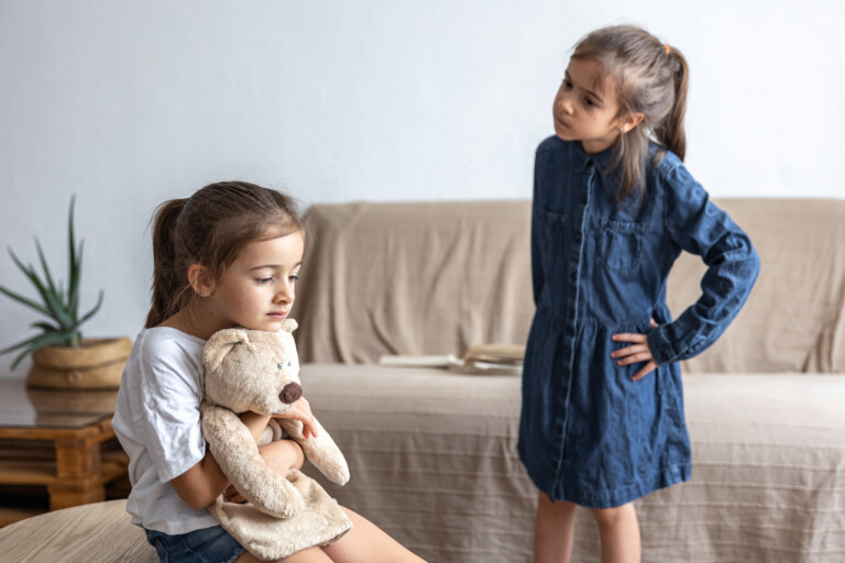 Dos hermanas pequeñas después de una pelea: una sentada abrazando su peluche y la otra de pie con las manos en la cintura