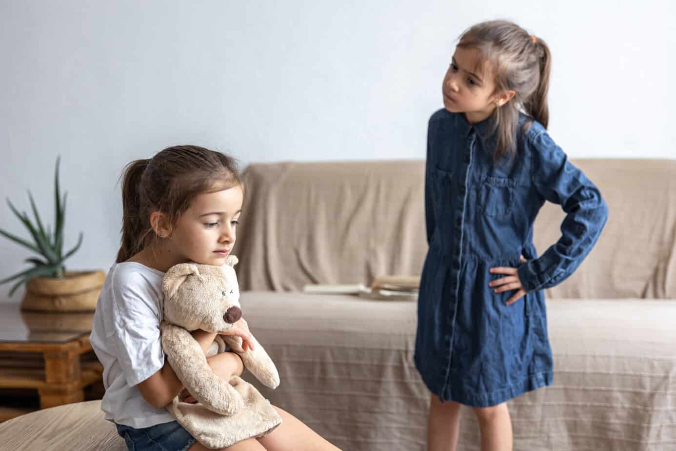Dos hermanas pequeñas después de una pelea: una sentada abrazando su peluche y la otra de pie con las manos en la cintura