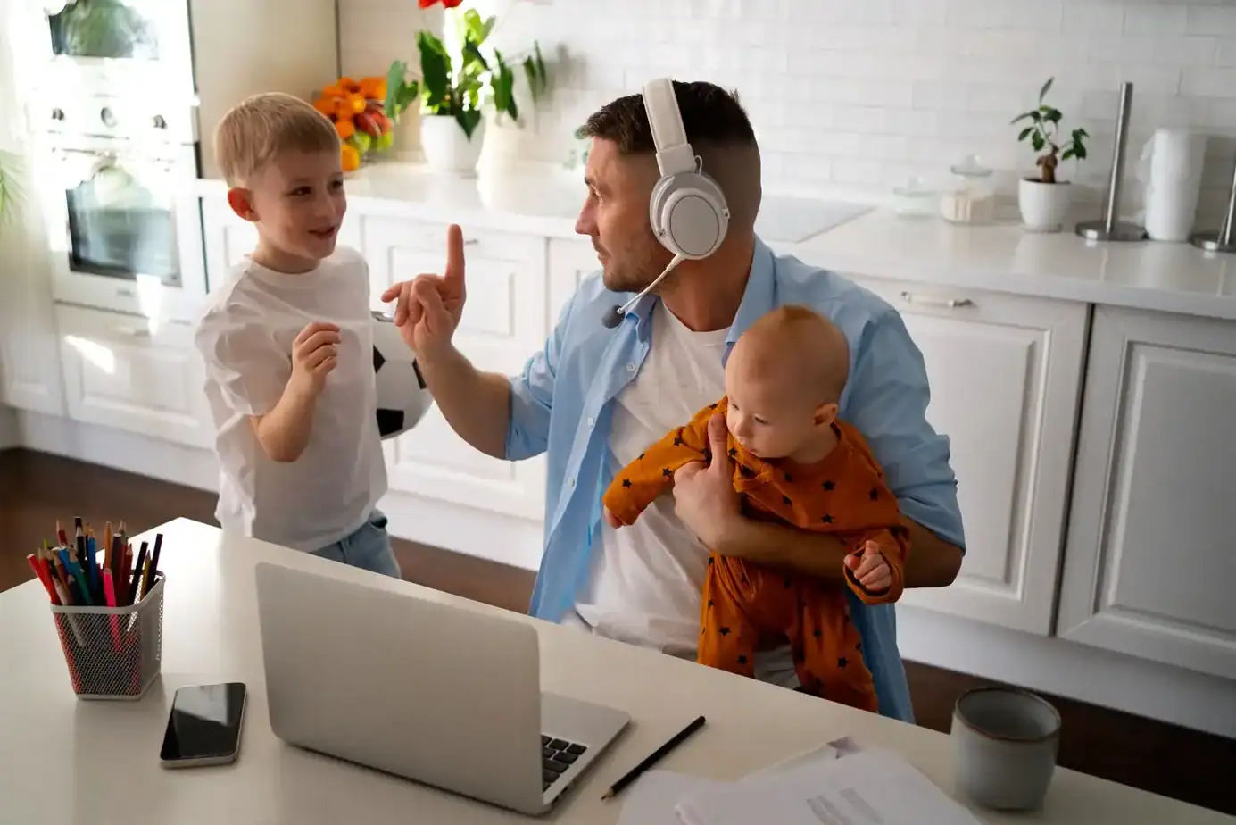 Teletrabajo con niños en casa: cómo crear un espacio saludable y organizar las rutinas familiares 1 Padre teletrabajando con auriculares mientras atiende a sus dos hijos pequeños en casa.